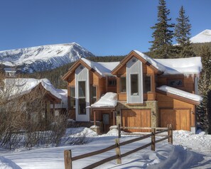 A wooden cabin with a snow-covered roof, surrounded by a wooden fence and snow-covered ground, with mountains in the background.