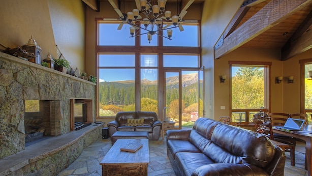 Ein geräumiges Wohnzimmer mit Kamin aus Stein, Ledersofas und einem großen Fenster mit Blick auf die Berge.