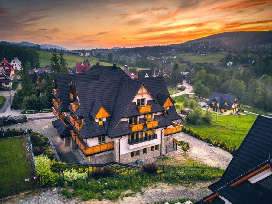 Un immeuble à plusieurs étages, doté d’un toit à forte pente, de nombreux balcons et entouré d’un paysage verdoyant avec des collines à l’horizon.
