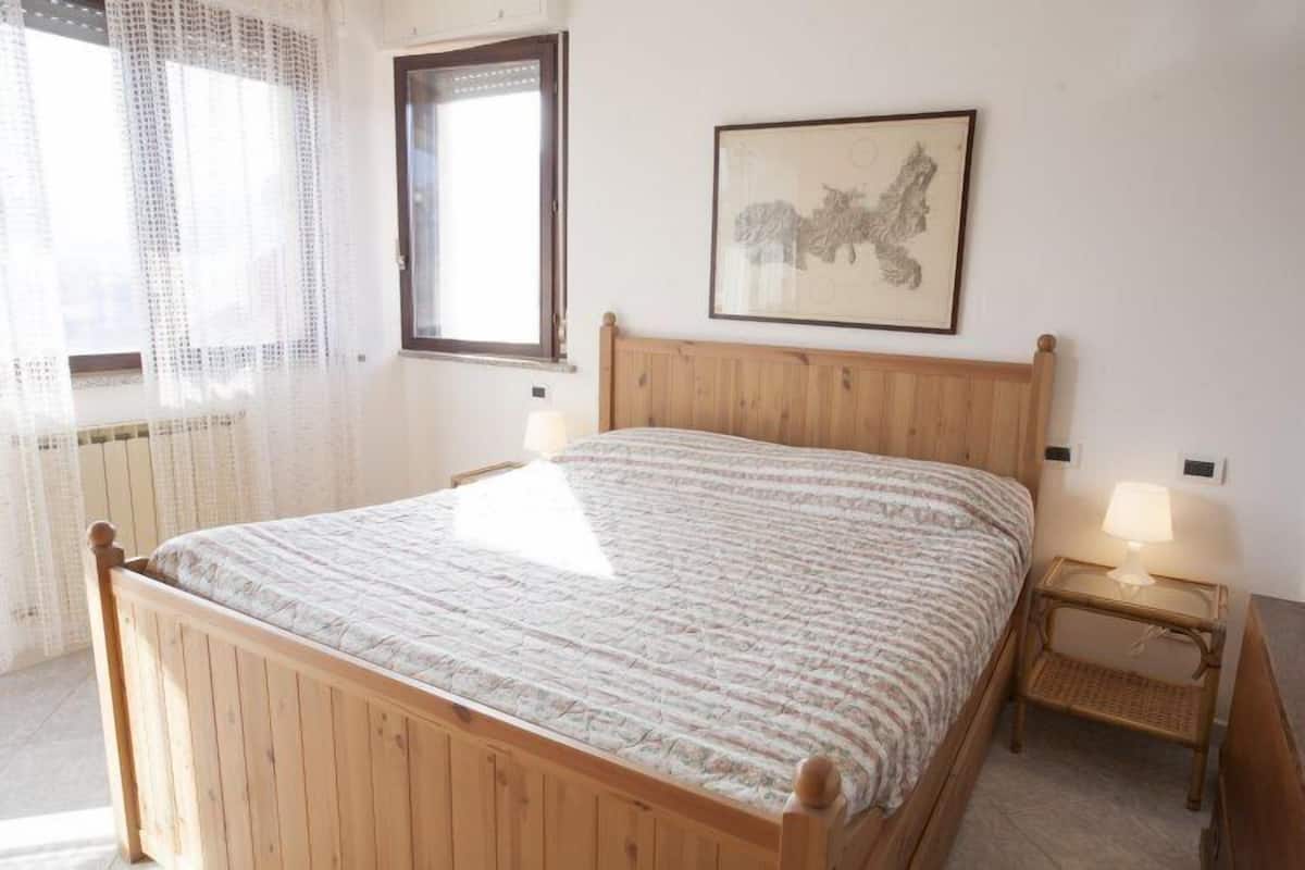 A bedroom with a wooden bed, a window with curtains, a bedside table with a lamp, and a framed picture on the wall.
