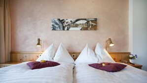 A double bed with white linens and purple pillows, a painting on the wall, bedside lamps, and a small table with a vase of flowers. - T3 Hotel Mira Val (Flims)