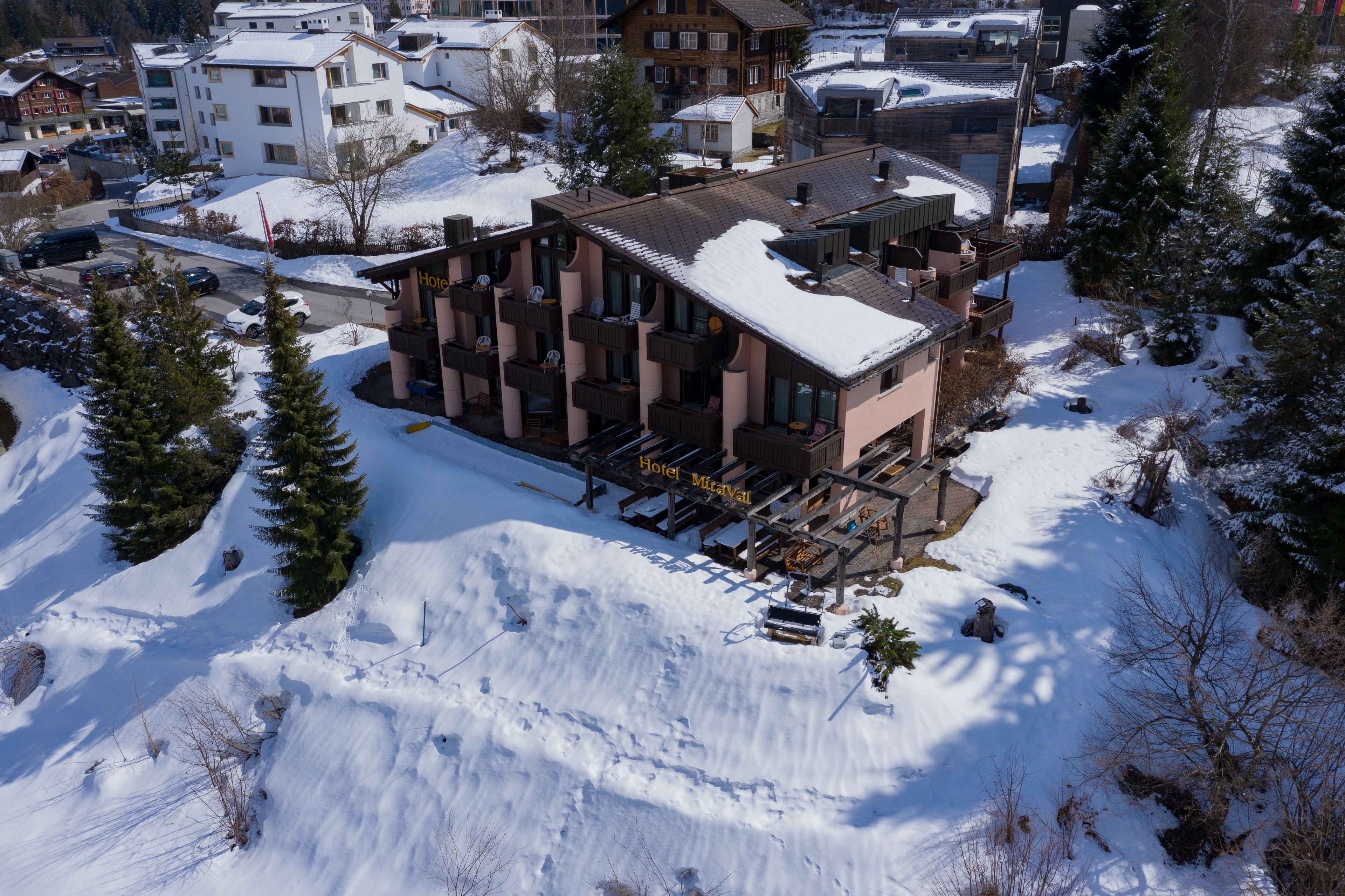 Ein Hotel mit verschneitem Dach, mehreren Balkonen und einer Skipiste im Hintergrund.