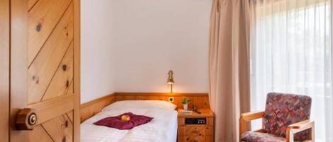 A wooden bed with a headboard, a bedside table with a lamp, a chair, and a window with curtains.