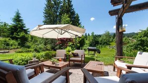 An outdoor patio with wooden furniture, a large umbrella, and a view of a lush garden and forest. - T3 Hotel Mira Val (Flims)