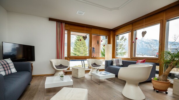 A modern living room with a sofa, chairs, a coffee table, and a TV. Large windows offer a view of mountains. - T3 Hotel Mira Val (Flims)