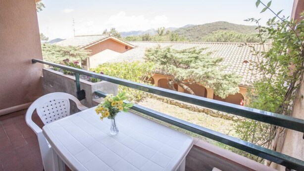 En balkon med et hvidt bord og stol, en vase med blomster og udsigt til en bygning og træer.