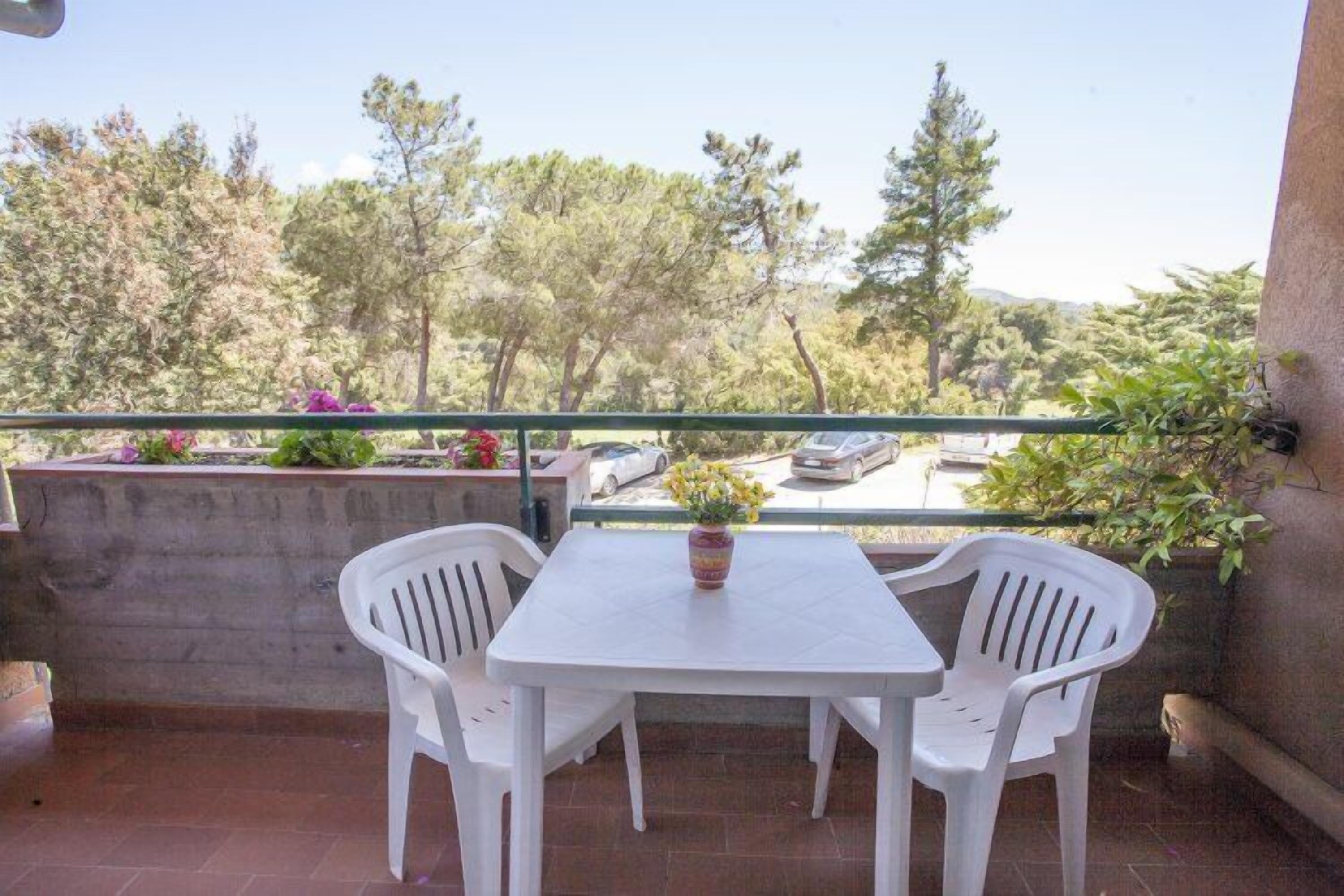 A balcony with a white table and two white chairs, overlooking a parking area and trees.