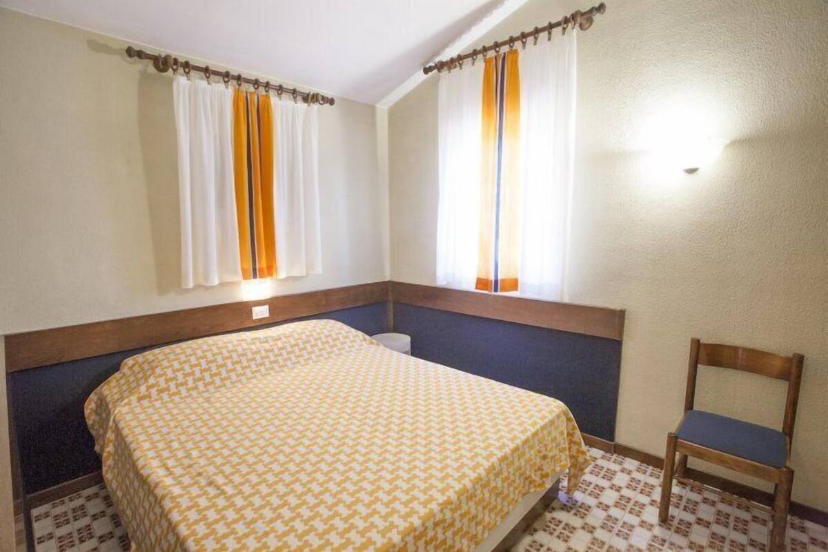 Quarto de hotel com cama, poltrona, janela com cortinas e luminária na parede.
