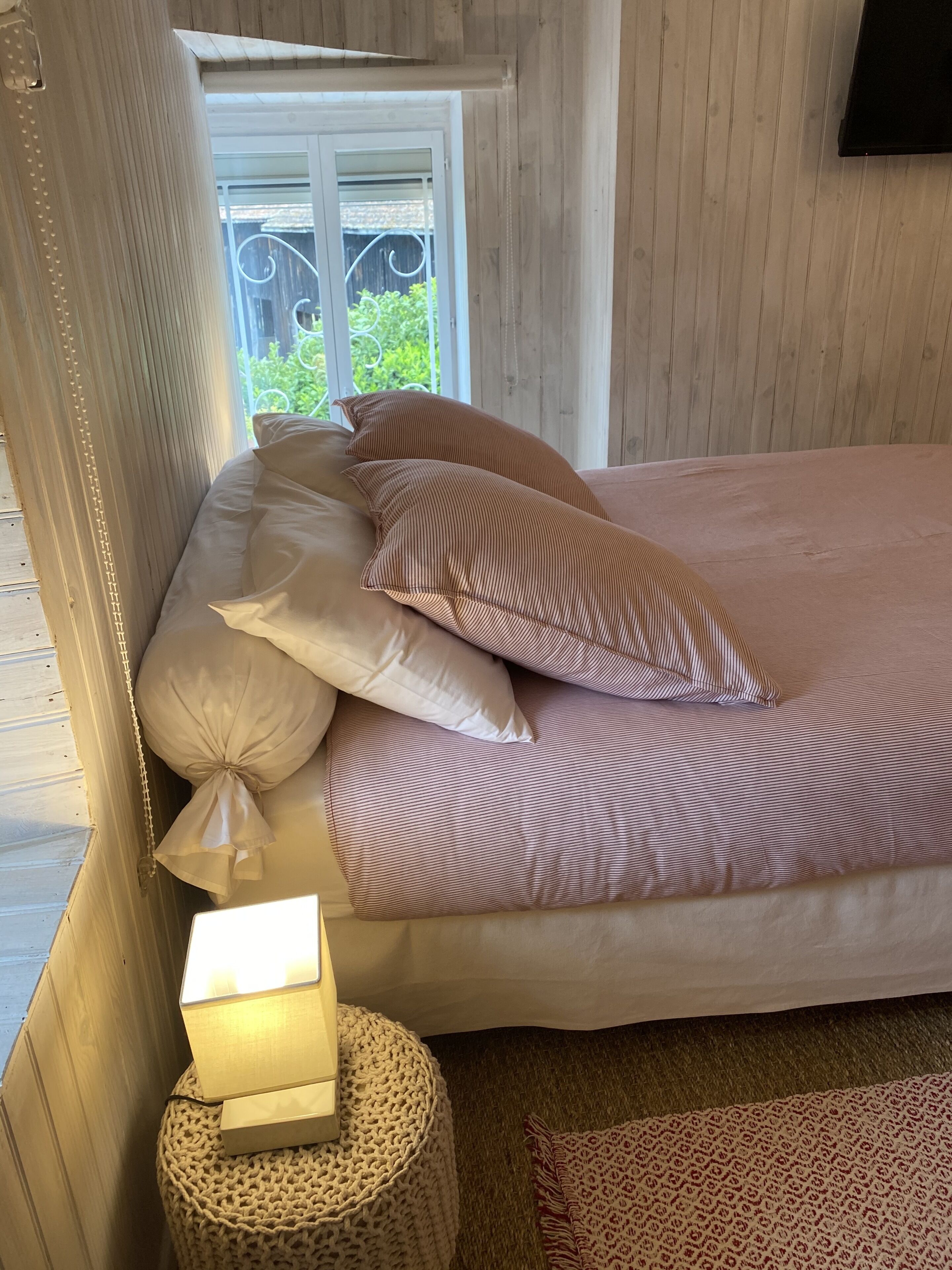Una cama bien hecha con ropa de cama a rayas, una lámpara de noche y un taburete tejido.