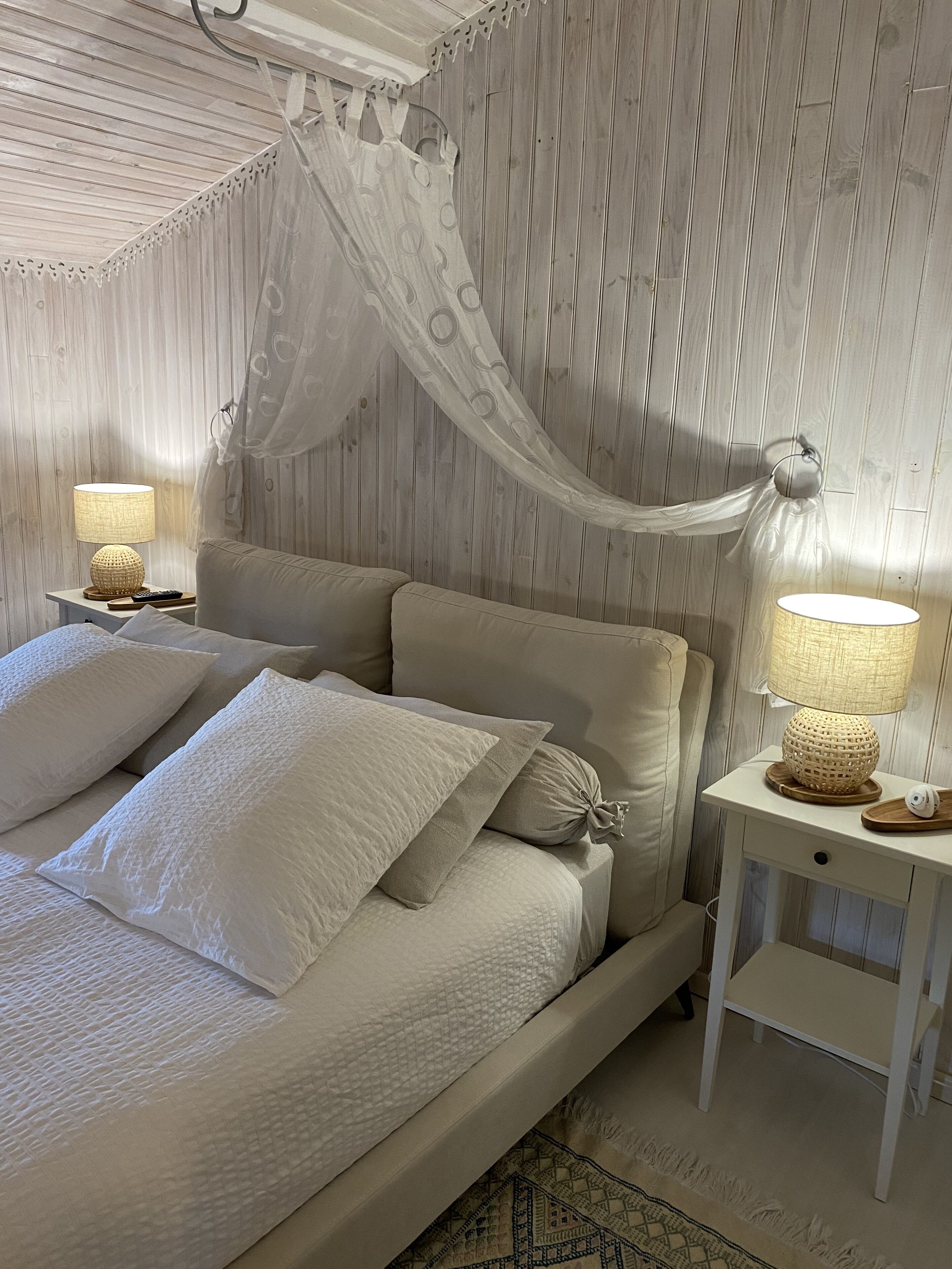 Un dormitorio con una cama, dos mesitas de noche, una lámpara, una cortina y una alfombra estampada.