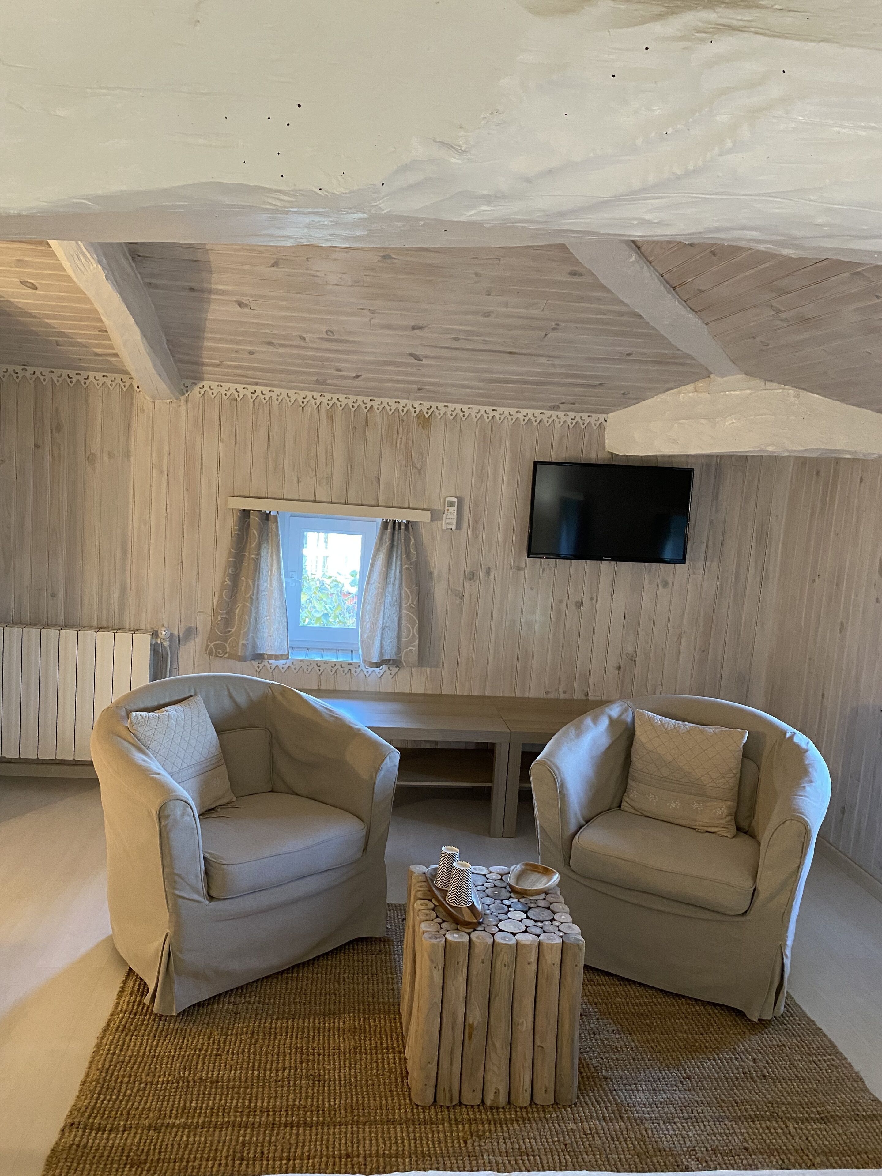 Een knus ingerichte kamer met houten wanden, een flatscreen televisie aan de wand, twee leren fauteuils, een houten bijzettafeltje en een raam met gordijnen.