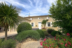 A stone building with a tiled roof, surrounded by a garden with various plants and flowers.