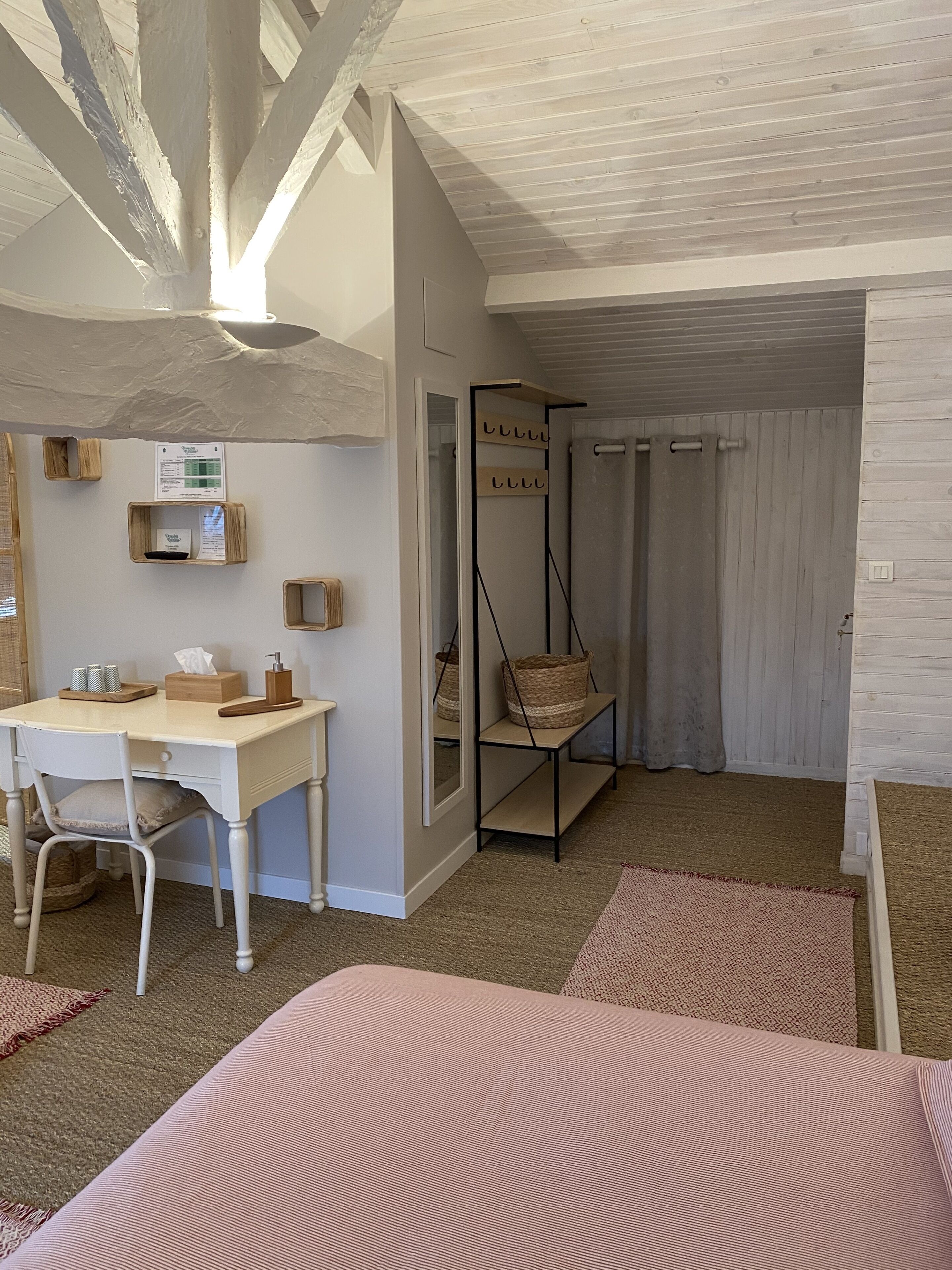 A room with a desk, chair, and a bed. There is a wooden shelf with baskets and a hanging basket. The room has a sloped ceiling and a door leading to another room.