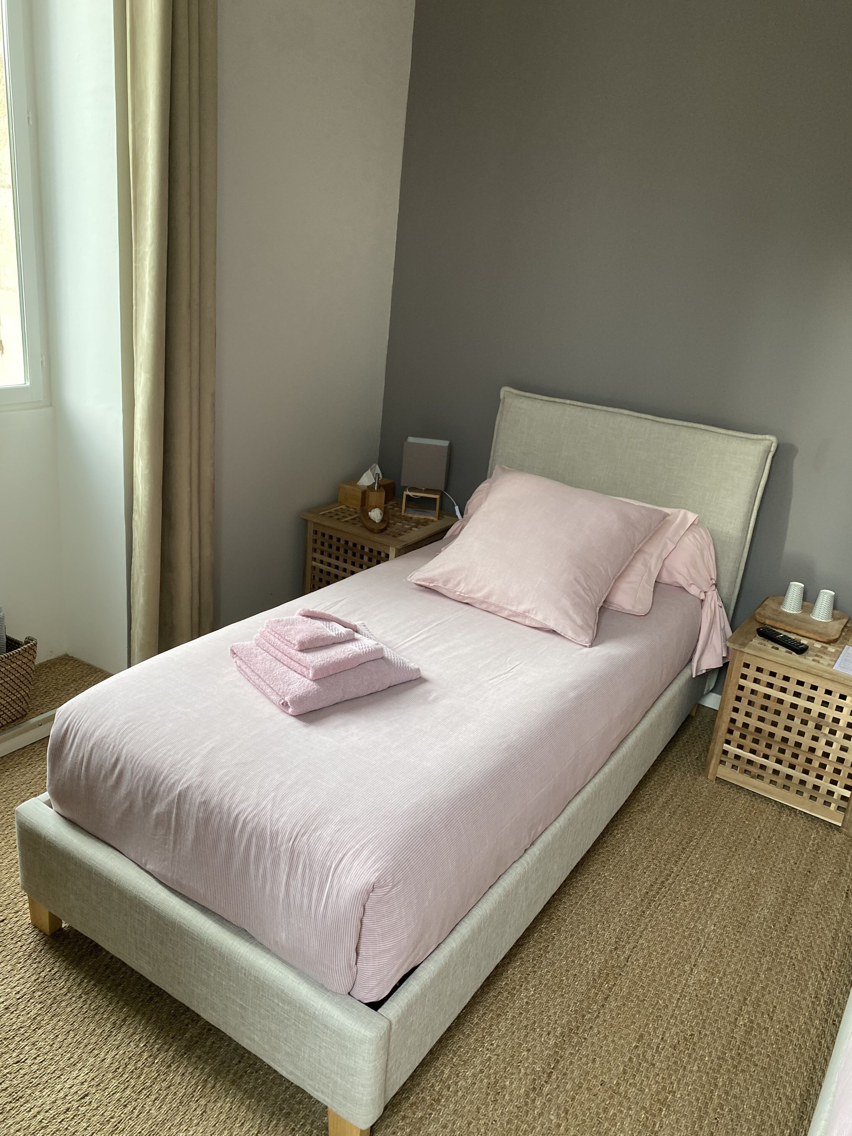 A neatly made bed with a light-colored headboard and footboard, a bedside table with a lamp and a tissue box, and a small wooden nightstand with a lamp and a tissue box.