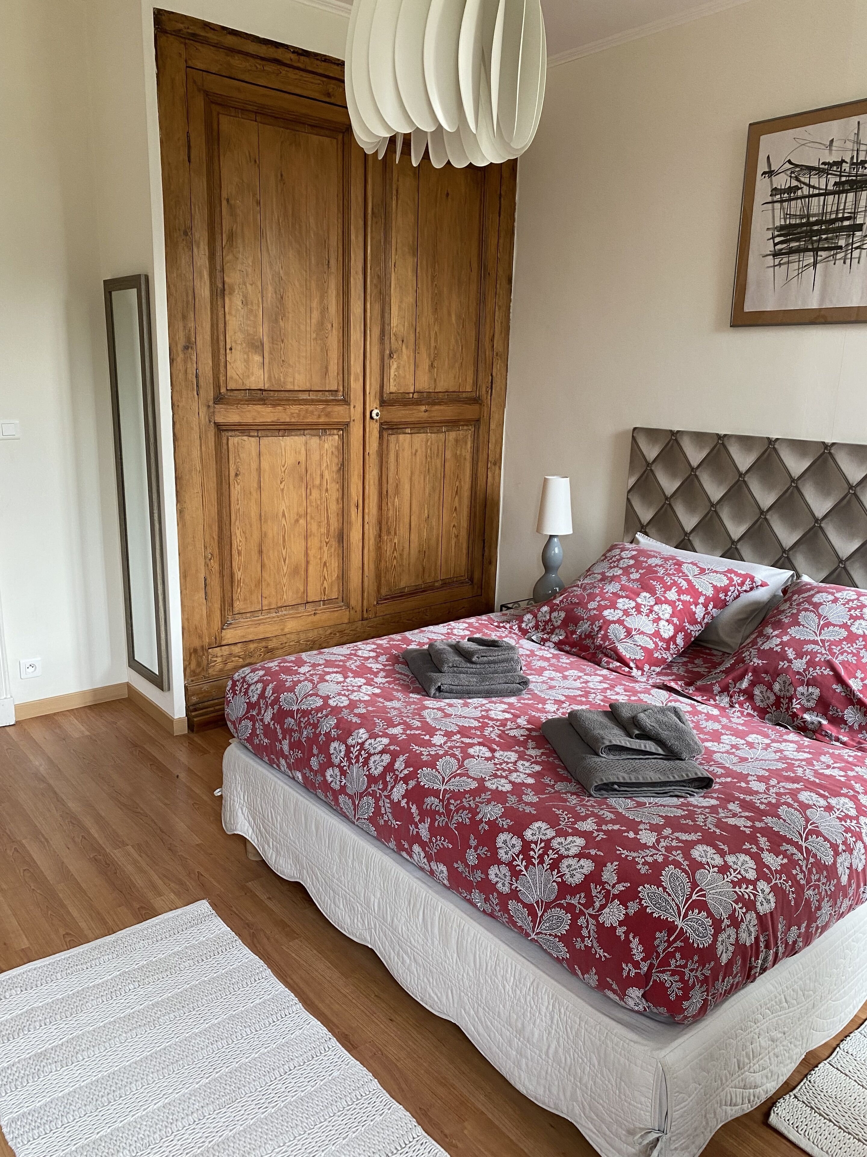 A bedroom with a wooden wardrobe, a bed with floral bedding, and a bedside table with a lamp.
