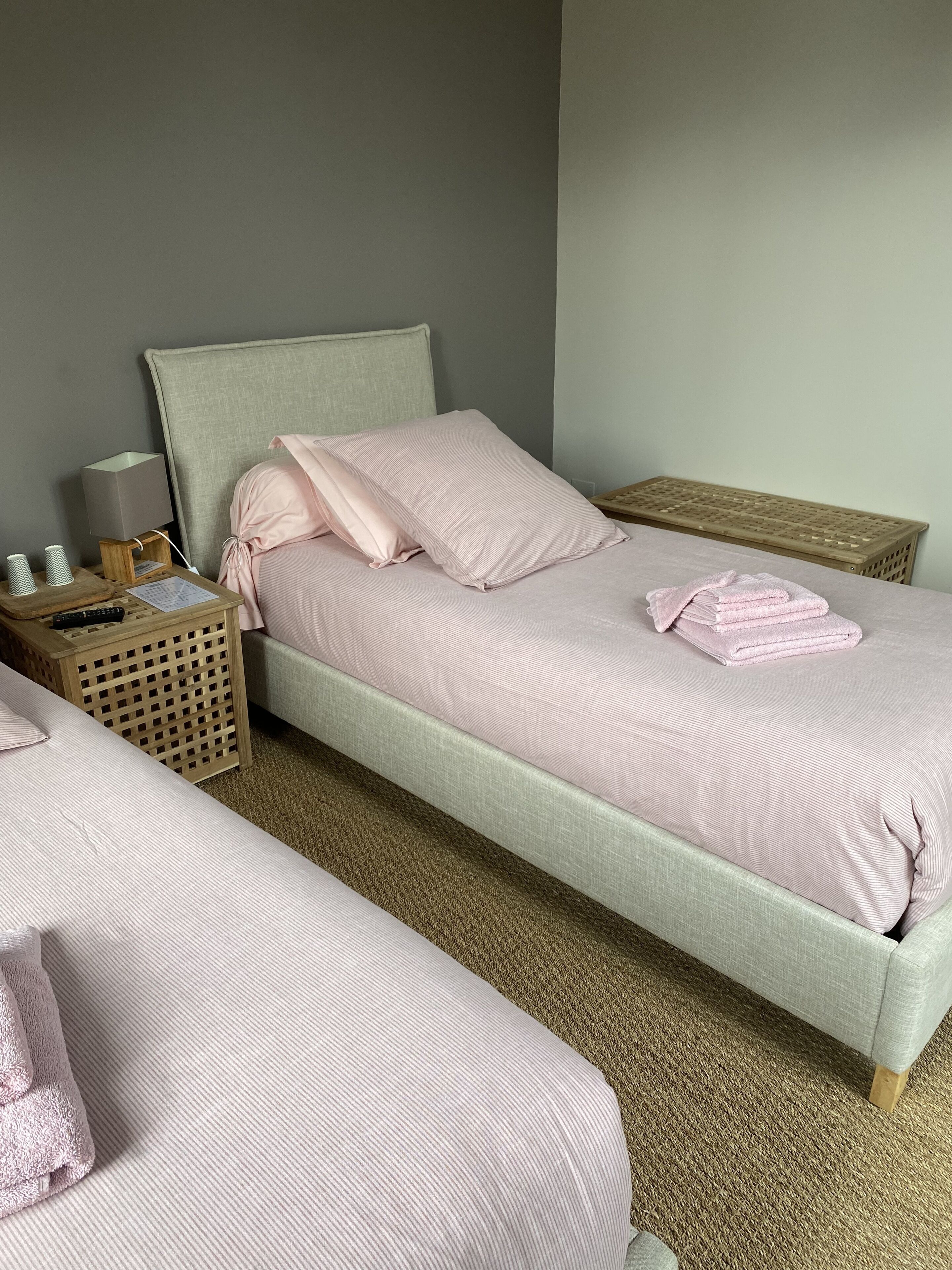 Habitación doble con dos camas, cada una con cabecero y piecero a juego, una mesita de noche con lámpara y una pequeña mesita auxiliar de madera.
