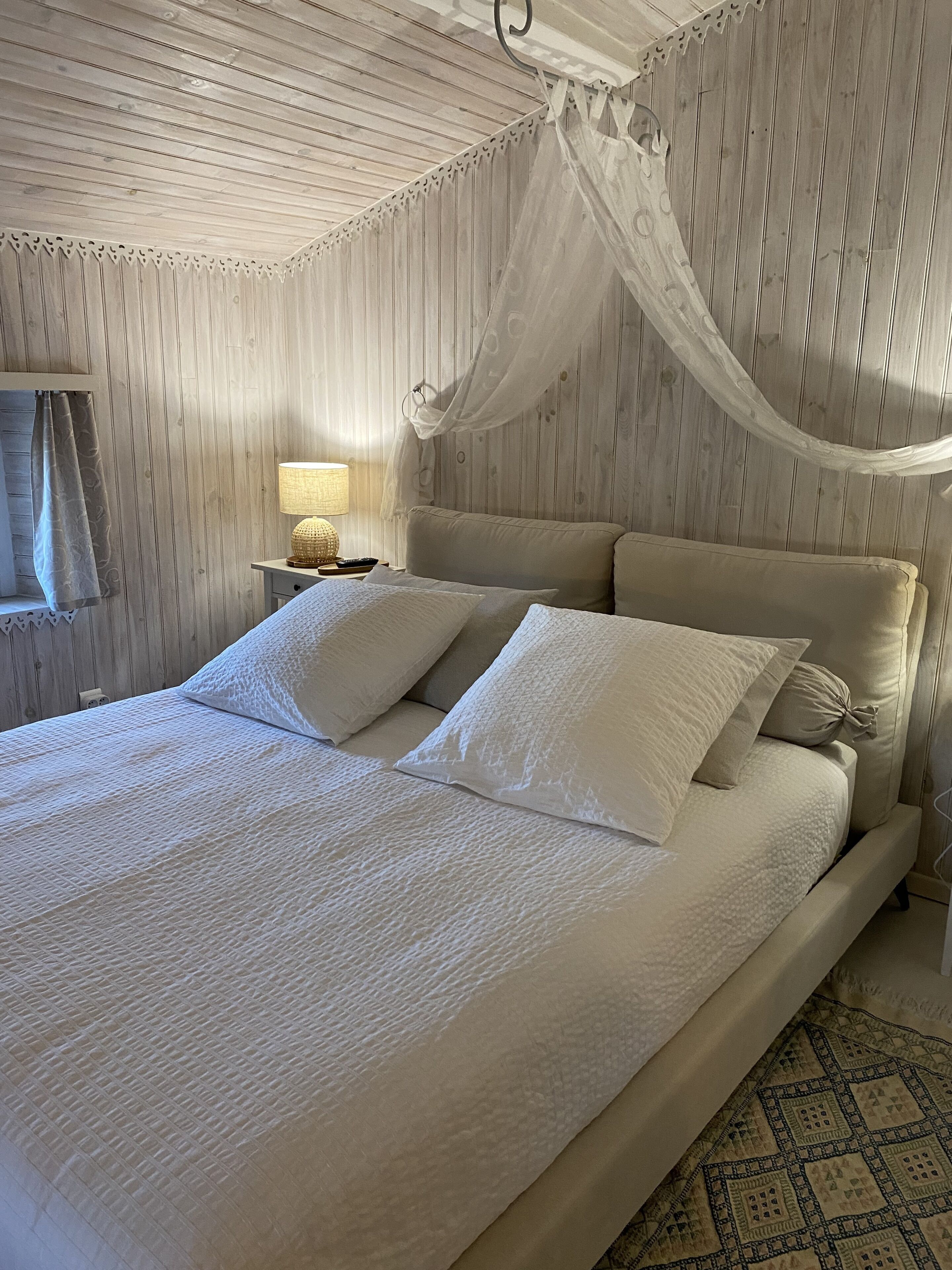 Una cama con ropa blanca y dosel, una mesita de noche con lámpara y pared con diseño.