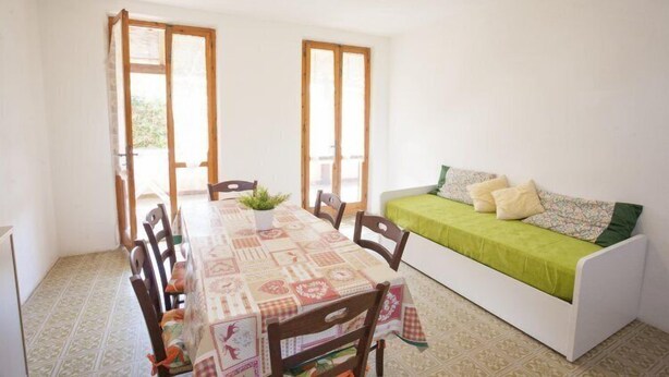 A small, bright room with a dining table, chairs, and a sofa. There are two windows with wooden frames and a door leading outside. - Appartamenti Le Mimose (Capoliveri)