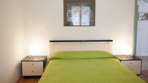 A bedroom with a bed, two bedside tables, a nightstand, a framed picture on the wall, and a door to the right. - Appartamenti Le Mimose (Capoliveri)