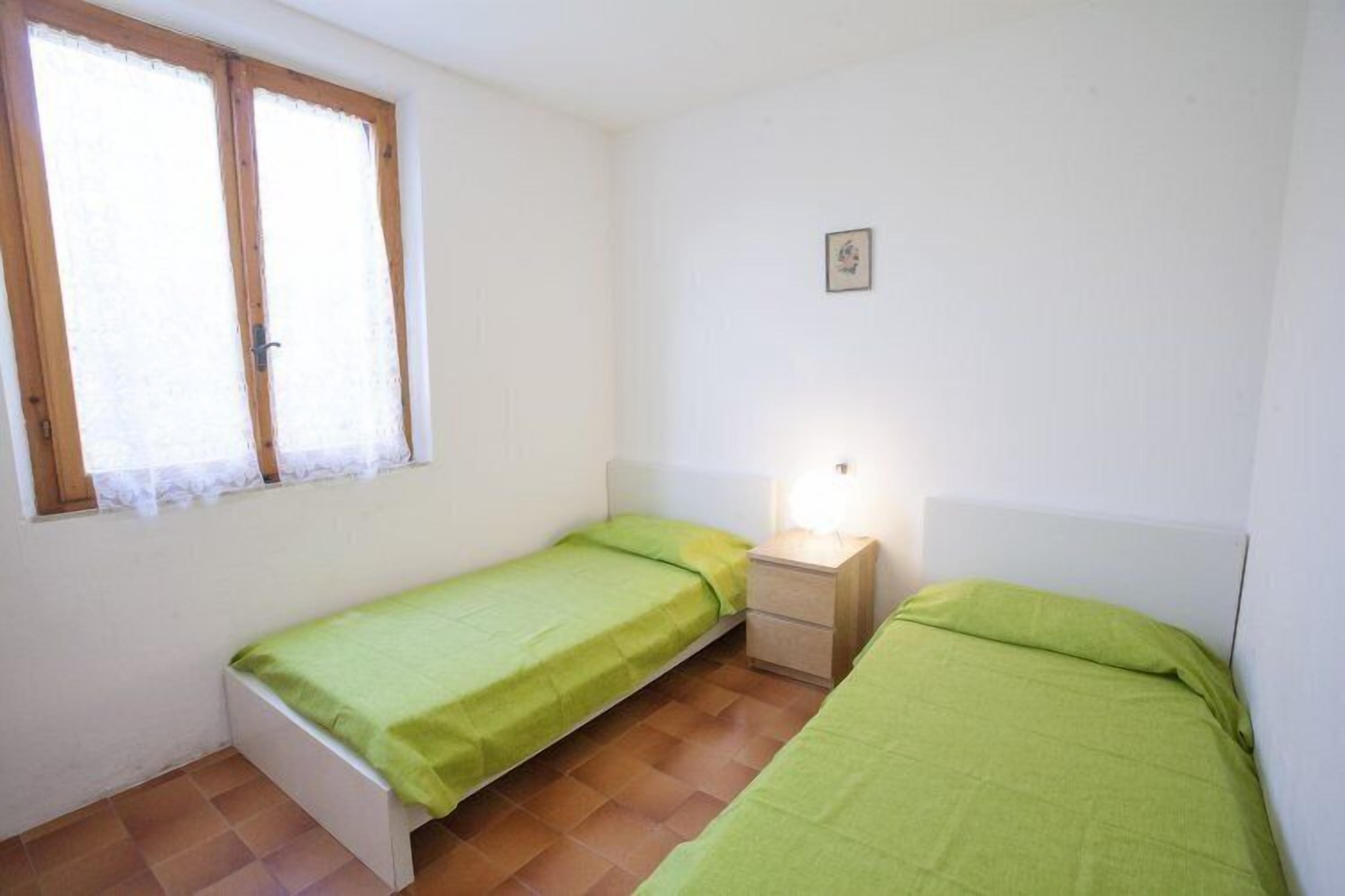 A small, clean room with two single beds, a window with curtains, a bedside table with a lamp, and a small picture on the wall.