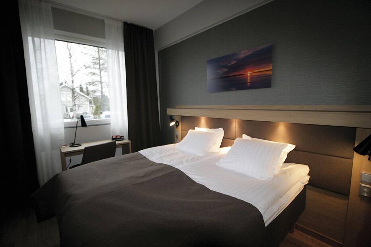 A hotel room with a large bed, a desk with a lamp, a window with curtains, and a painting on the wall.