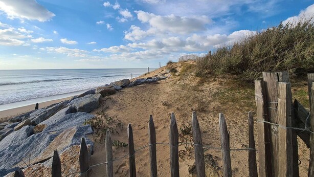 Une plage avec une clôture en bois et des rochers au premier plan, qui mènent à l’océan.