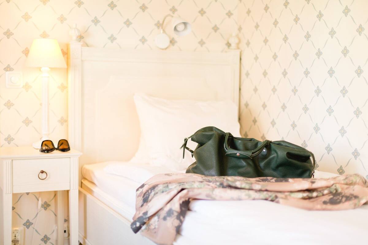 A neatly made bed with a green bag and sunglasses on the nightstand.