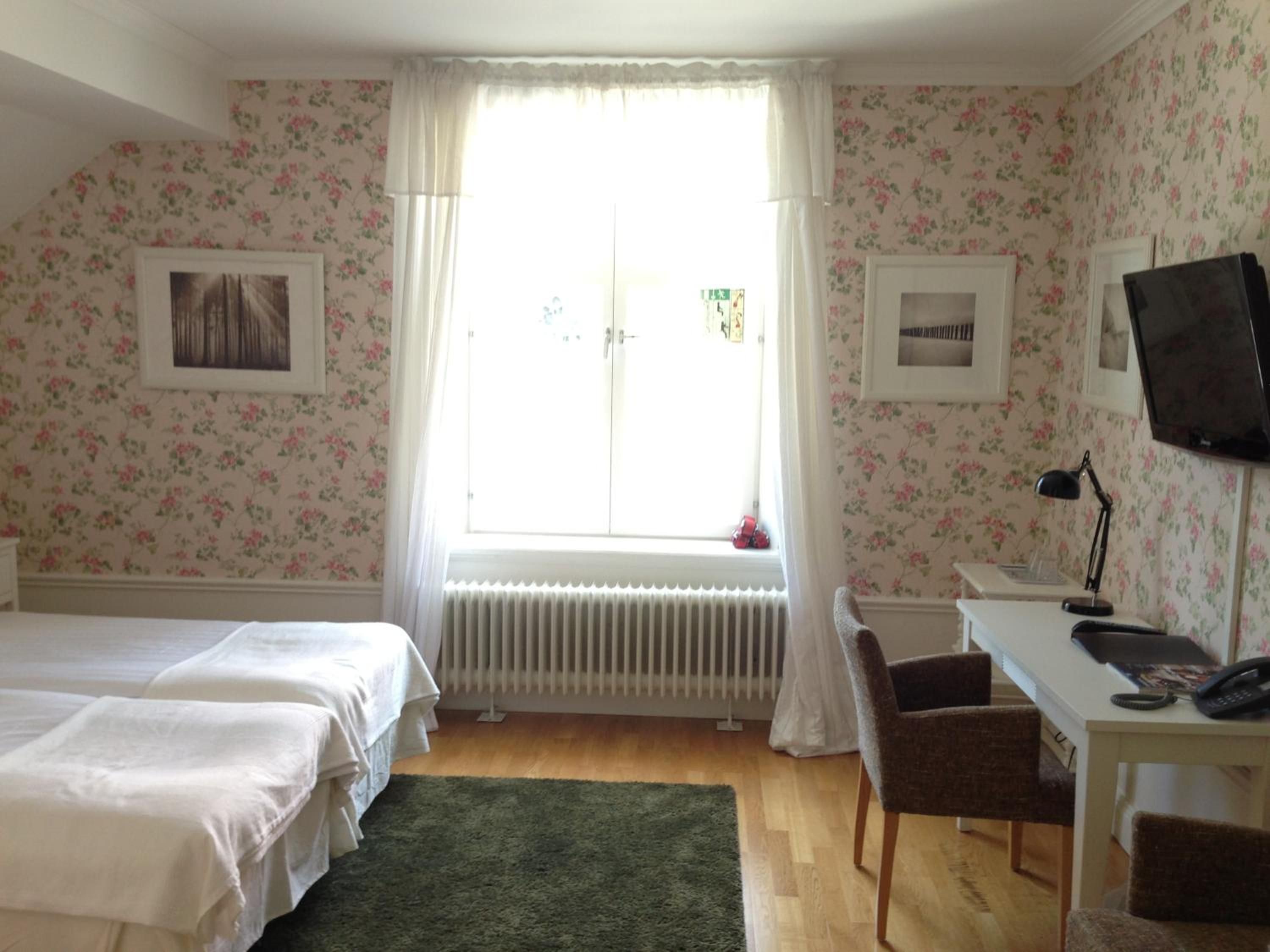 Quarto com duas camas, uma escrivaninha com cadeira, radiador e televisão.