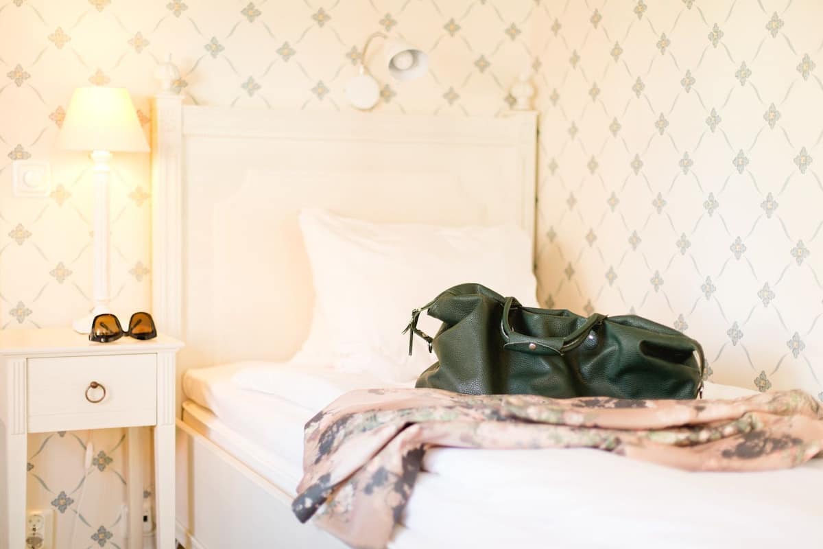 A neatly made bed with a green bag and sunglasses on the nightstand.