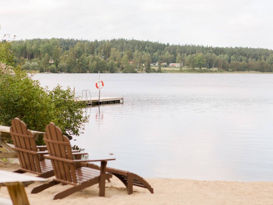 A lakeside setting with wooden chairs and a table, a dock extending into the water, and a forested hill in the background.