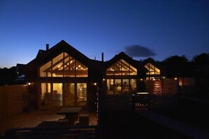 A modern house with large glass windows and a well-lit interior at dusk. - Merlin Farm Cottages Mawgan Porth (Newquay)