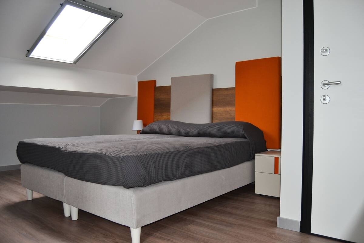 A modern bedroom with a sloped ceiling, a bed with grey bedding, an orange accent wall, and a bedside table with a lamp.