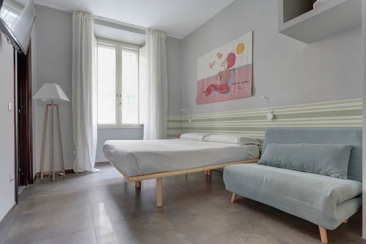Un dormitorio con cama, sofá, un cuadro en la pared y ventana con cortinas.