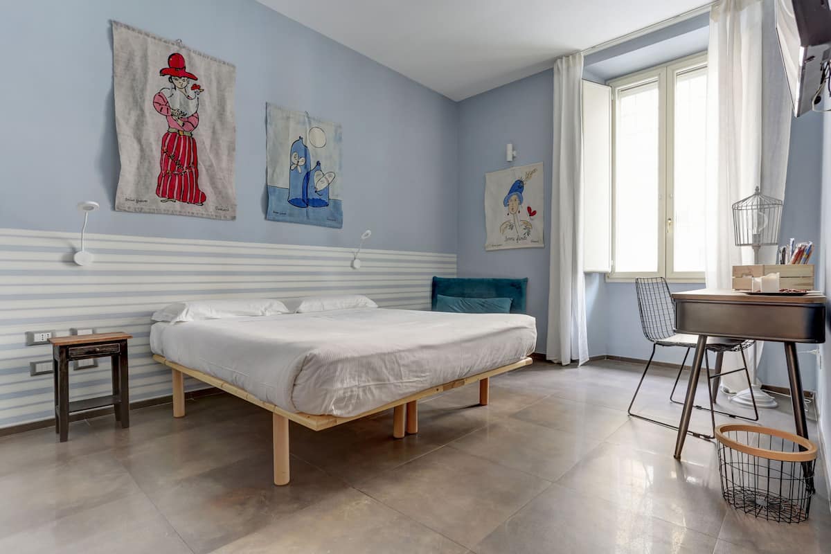 Un dormitorio con cama, escritorio y silla. Hay cortinas en la ventana y una obra de arte en la pared.