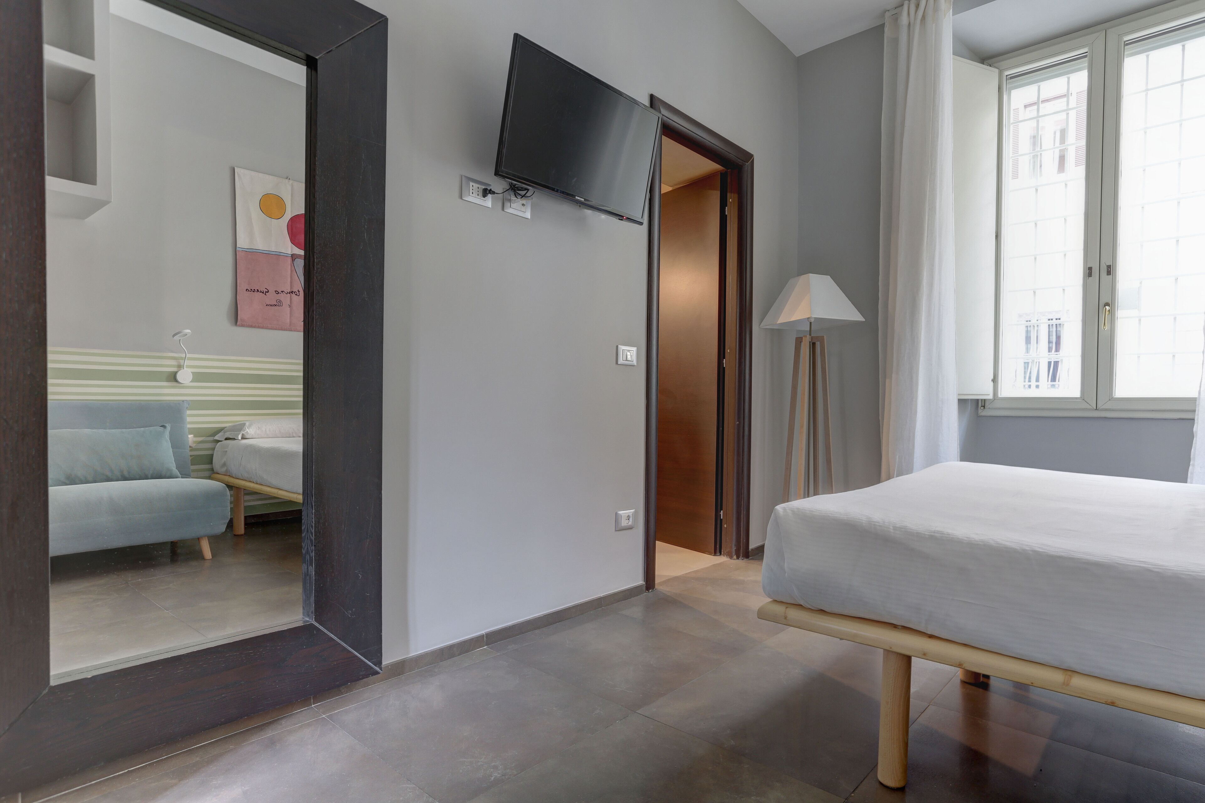 Habitación de hotel con cama, televisión montada en la pared, mesita de noche, lámpara y ventana con cortinas.