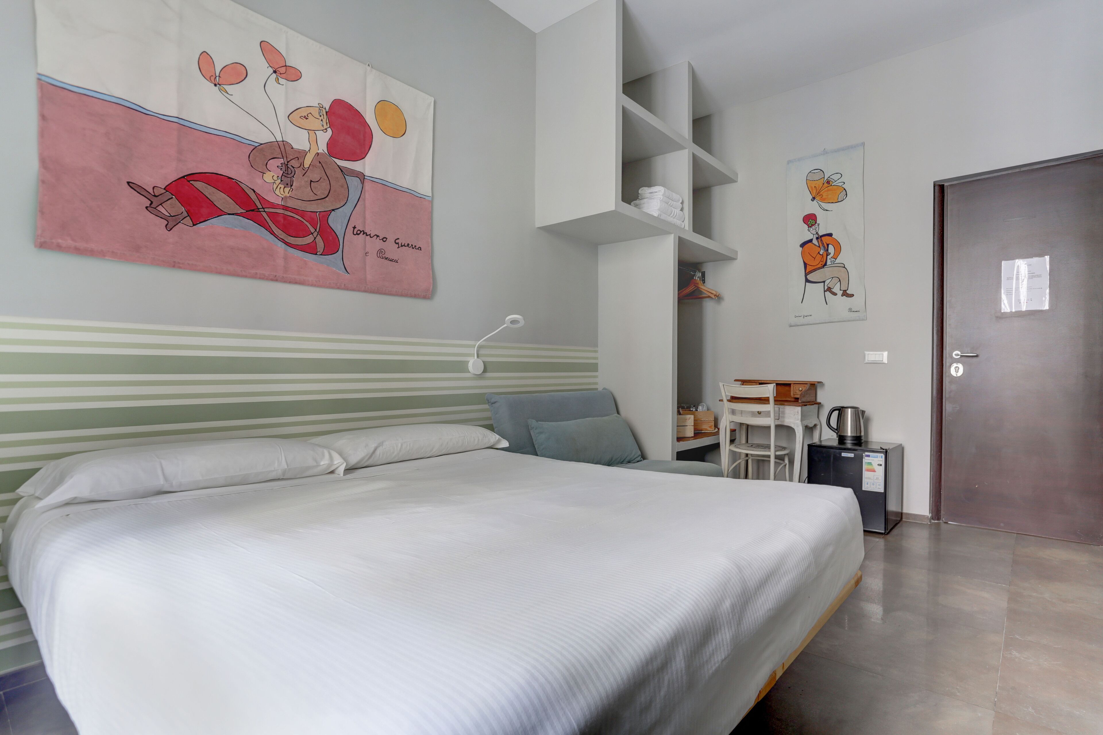 Un dormitorio con una cama, una mesita de noche, una silla, una cafetera y una puerta.