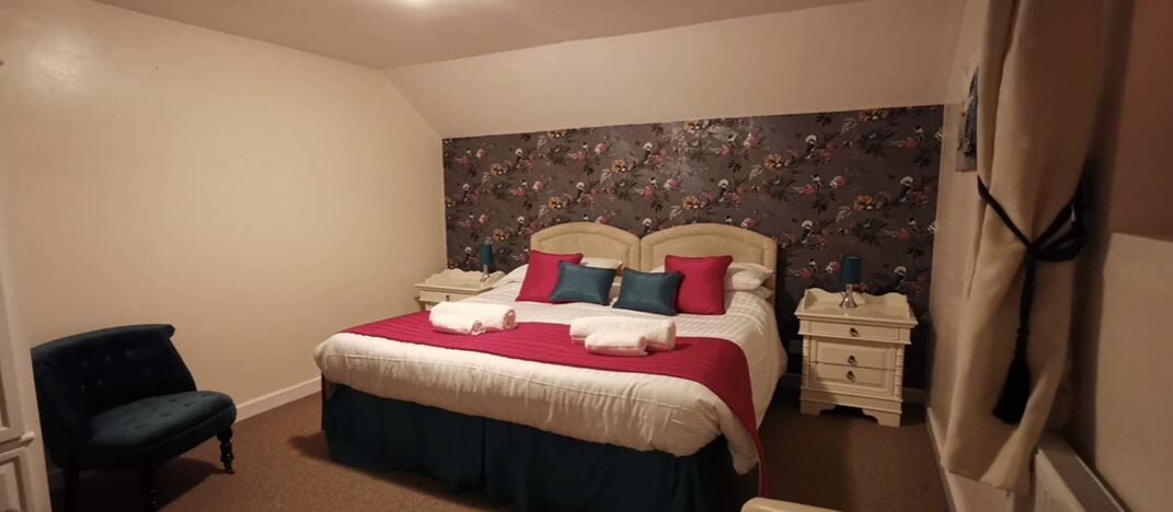 Un dormitorio con una cama, dos mesitas de noche, una silla y un papel tapiz con motivos florales.