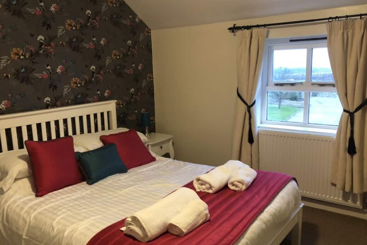 A neatly made bed with red and white bedding, a window with curtains, and a floral wallpaper pattern on the wall.