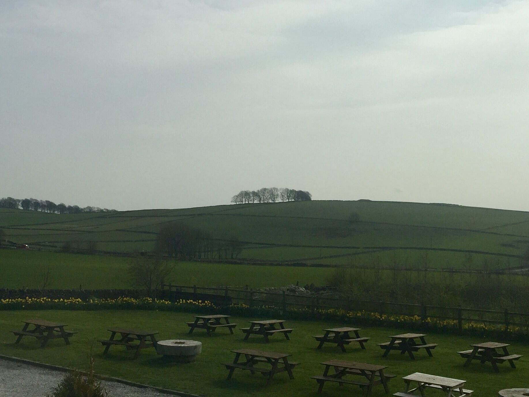 A rural landscape with rolling hills, a wooden fence, and picnic tables arranged in the foreground.