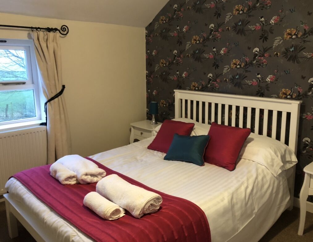 A neatly made bed with white linens and red and blue pillows, a red throw blanket, and two rolled towels on the bed.