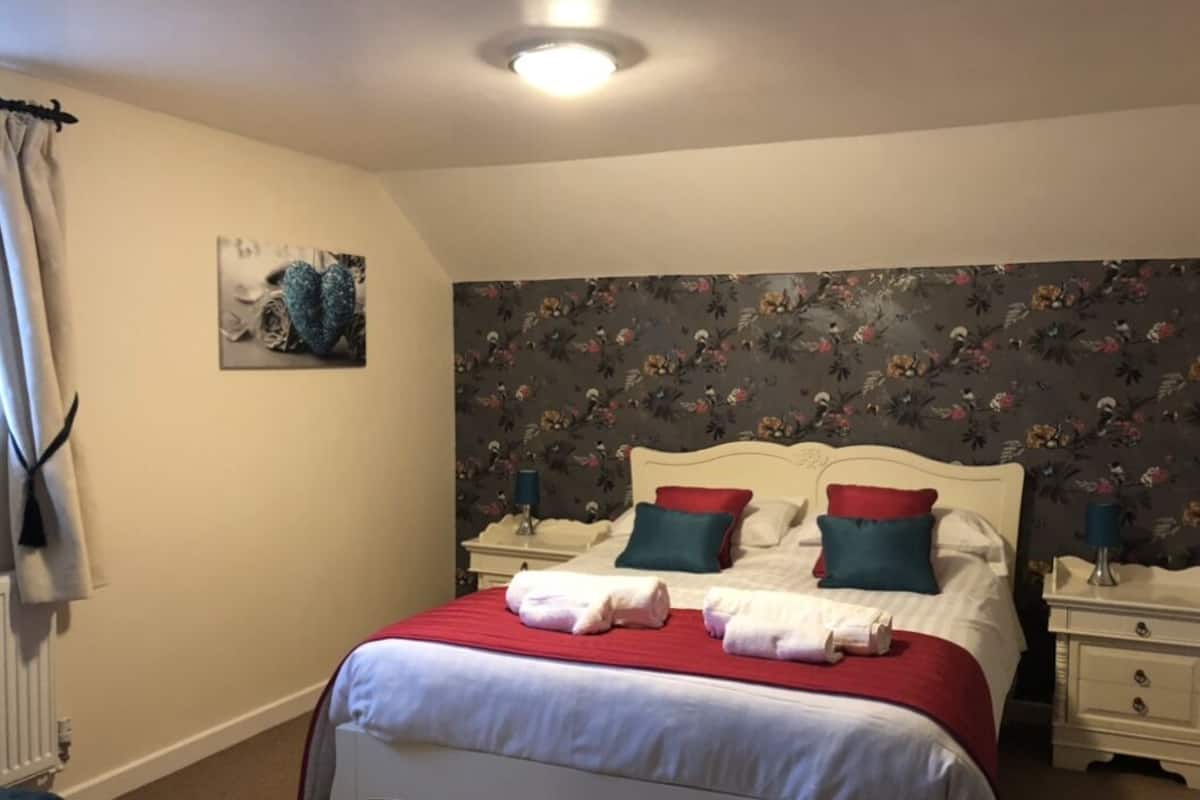 A double bed with red and white bedding, two bedside tables with lamps, a floral wallpaper, and a ceiling light.