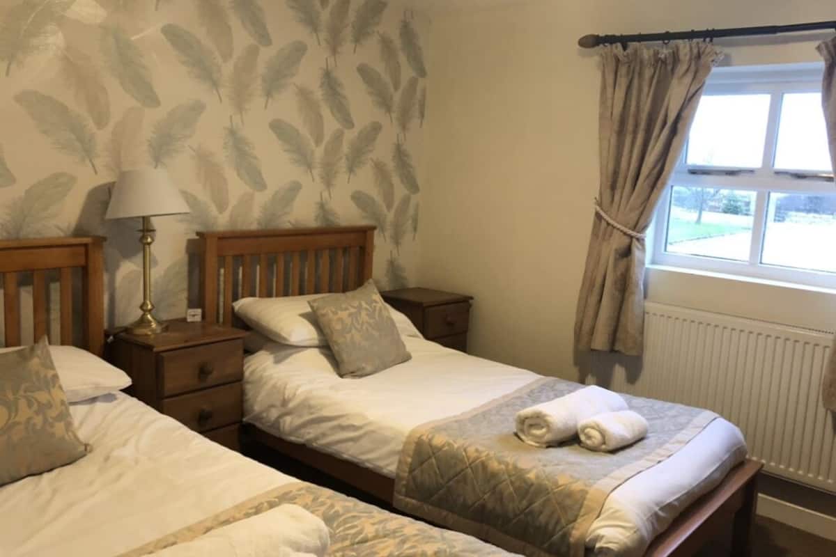 A hotel room with two beds, a nightstand with a lamp, a window with curtains, and a radiator.