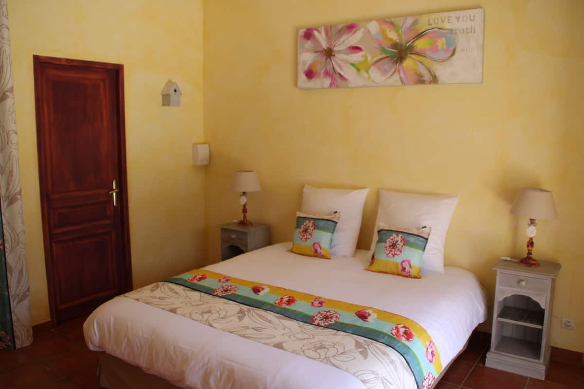 Une chambre à coucher comprenant un lit, deux tables de chevet, une commode, une porte en bois et un mur orné d’un tableau représentant des motifs floraux.