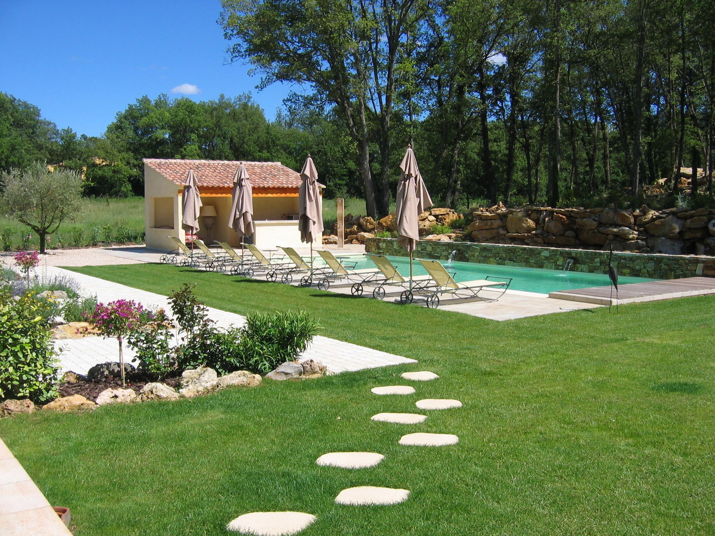 A pool area with lounge chairs and umbrellas, surrounded by a well-maintained lawn and a stone wall.