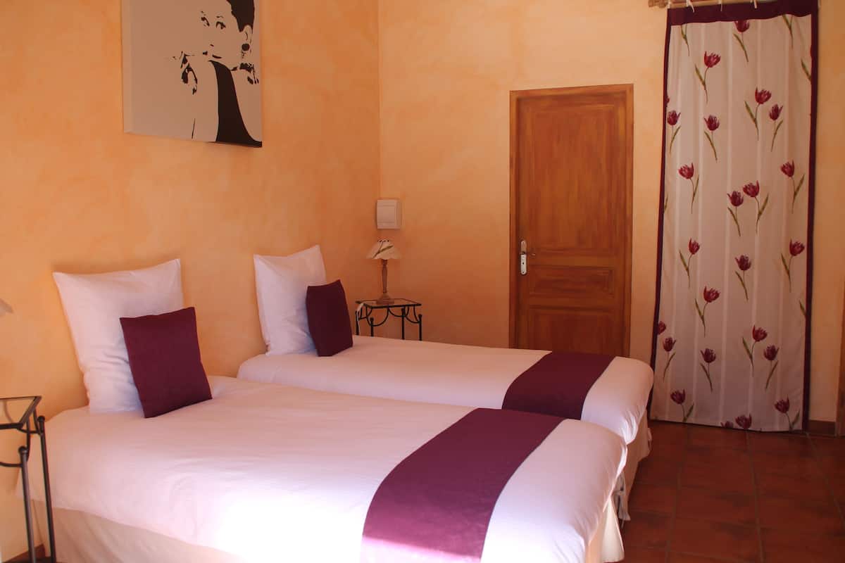 Une chambre d’hôtel avec deux lits, une porte en bois, une petite table avec une lampe et un rideau à motifs floraux.