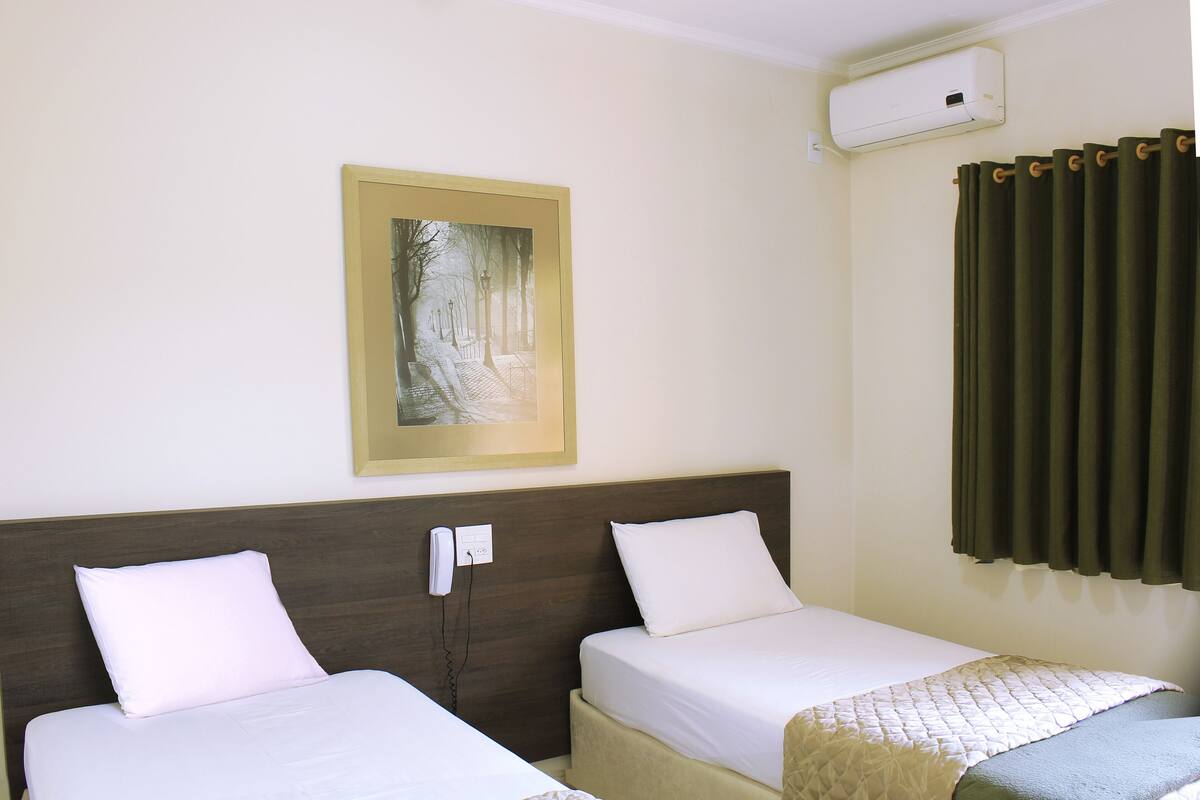 Une chambre d’hôtel avec deux lits, un climatiseur fixé au mur, un tableau encadré et une fenêtre avec des rideaux.