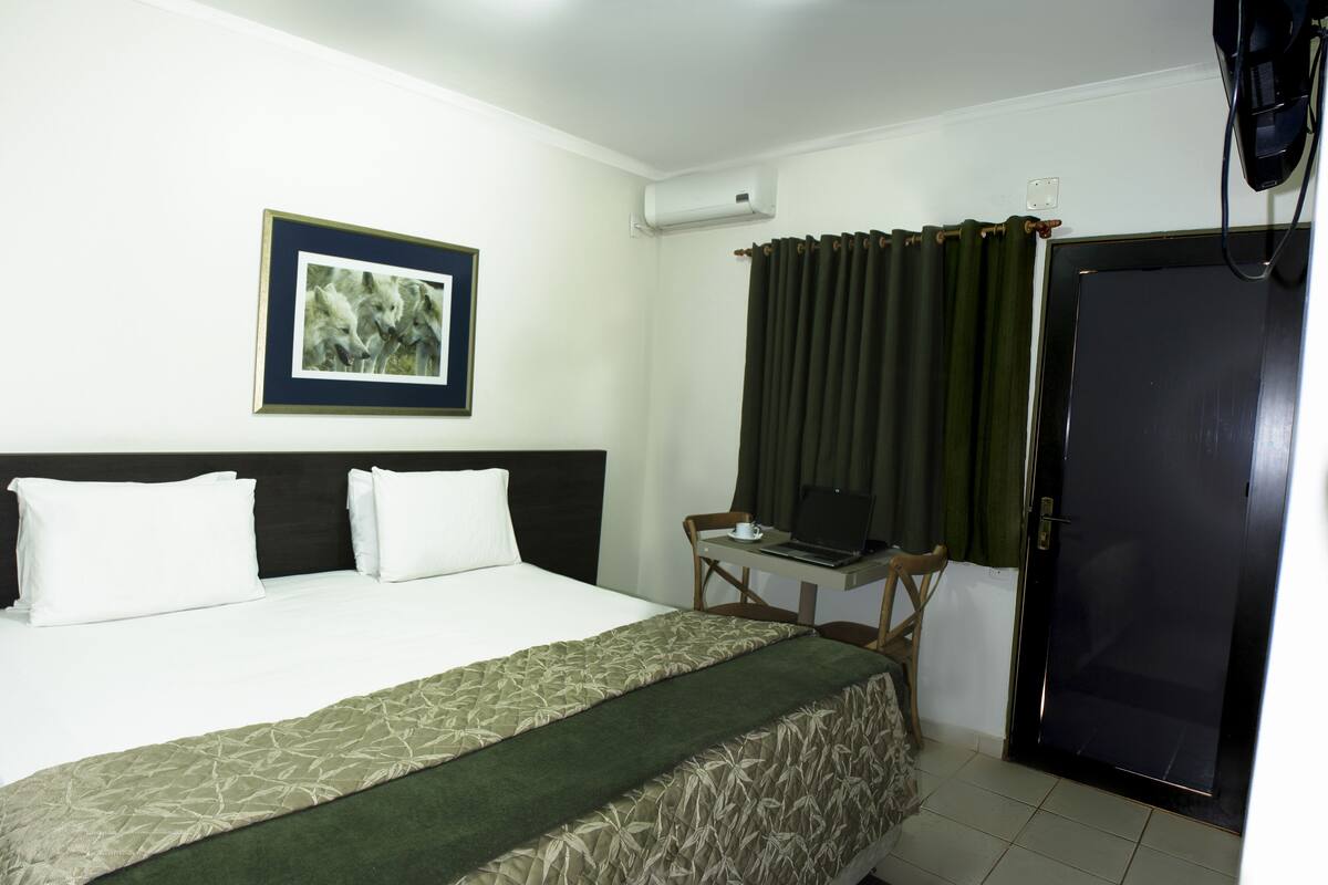 Une chambre d’hôtel avec un lit, un bureau avec un ordinateur portable, un tableau encadré au mur et une porte avec des rideaux.