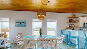 A dining area with a wooden ceiling, white chairs, a table with a woven light fixture, and a view of the outside through large windows. - Close to Beach, Pet Friendly and Shared Pool (Port Aransas)