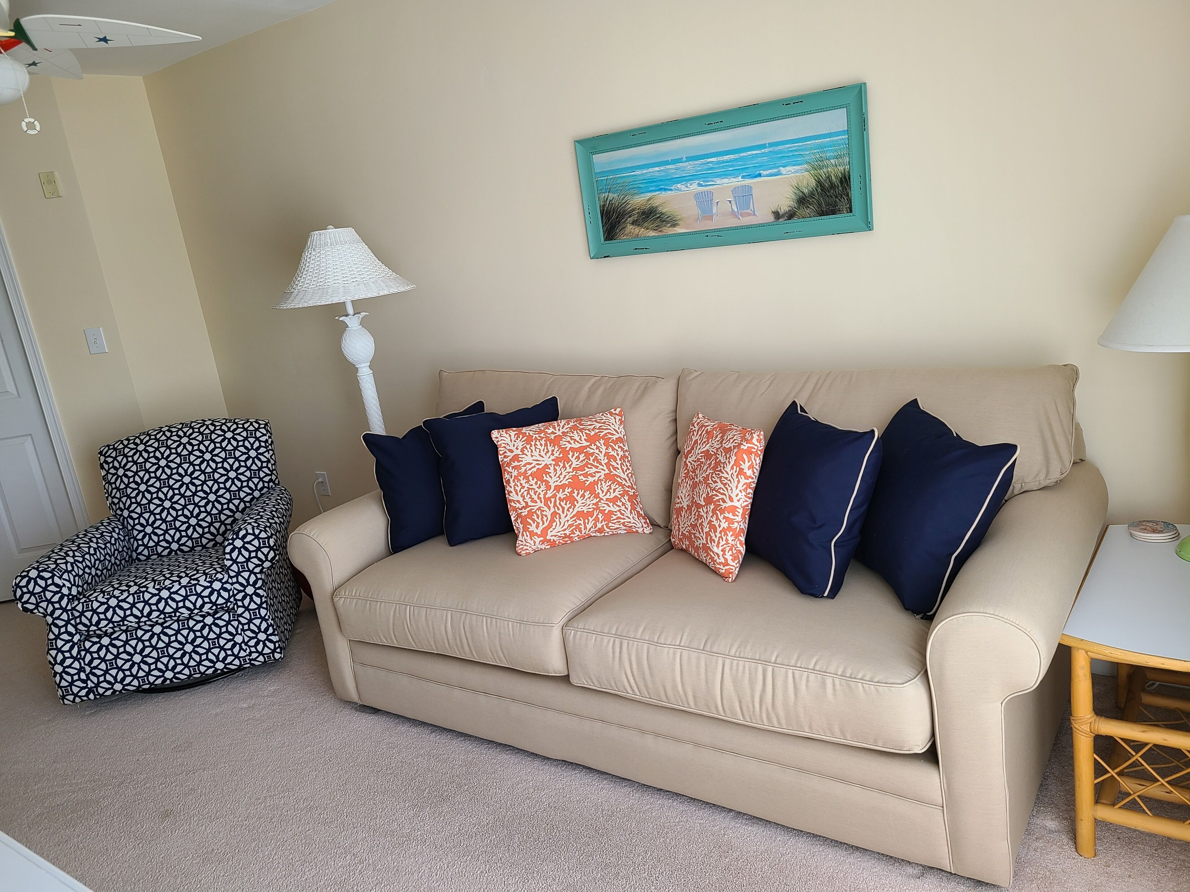 A beige sofa with multiple pillows, a patterned armchair, a small table with a lamp, and a framed picture on the wall.