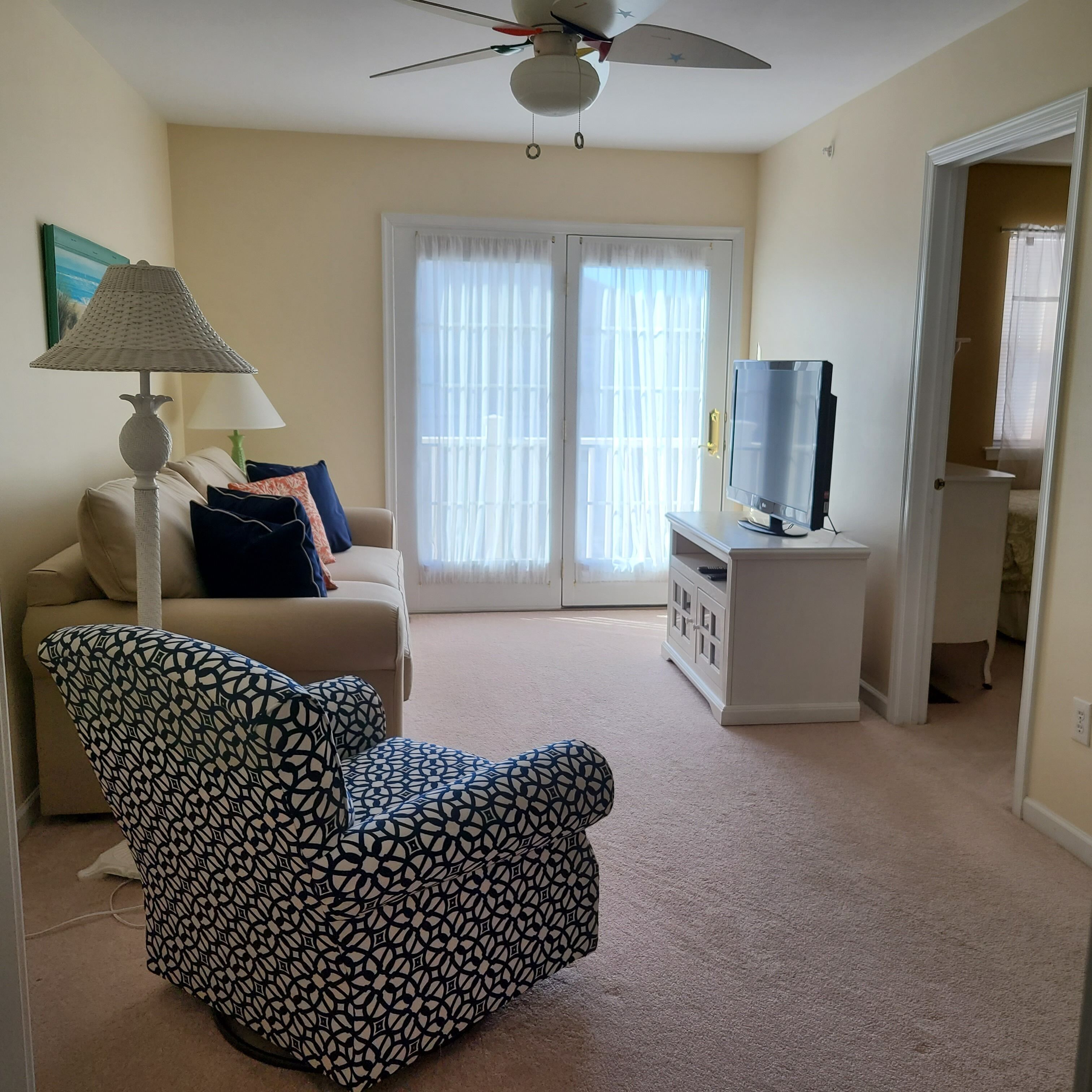 A living room with a patterned armchair, a lamp, a television on a stand, and a sliding glass door leading to a balcony.